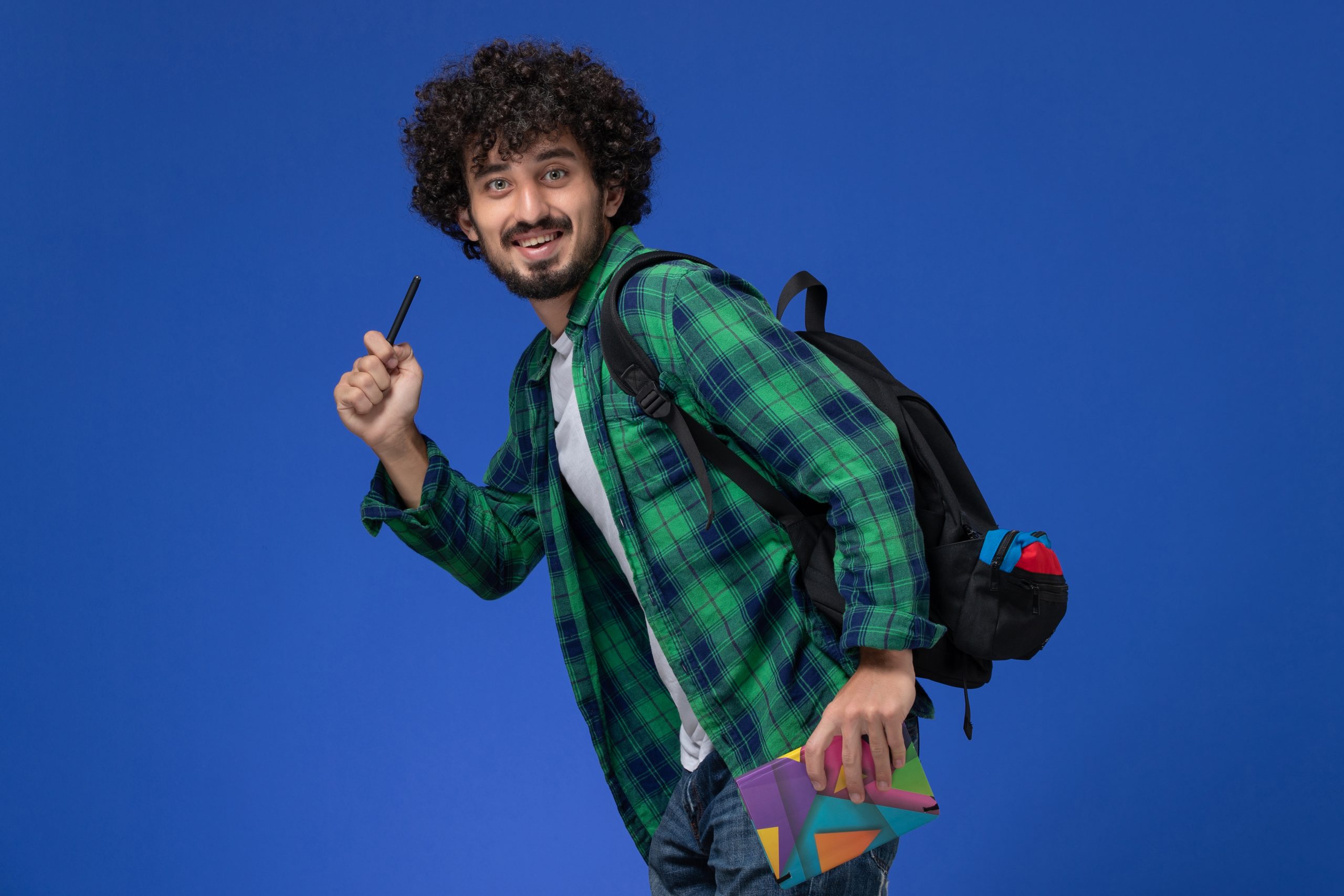 Estudante sorridente com mochila nas costas, caderno em uma mão e caneta na outra em posição de saída